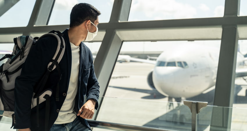 A man with a face covering and a backpack looks out an airport window at a plane