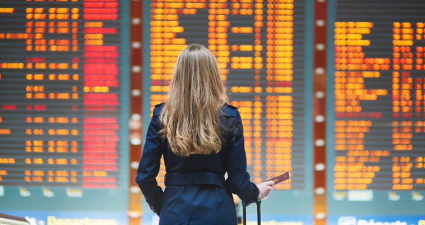 A woman with a suitcase stands in front of a large screen filled with flight information