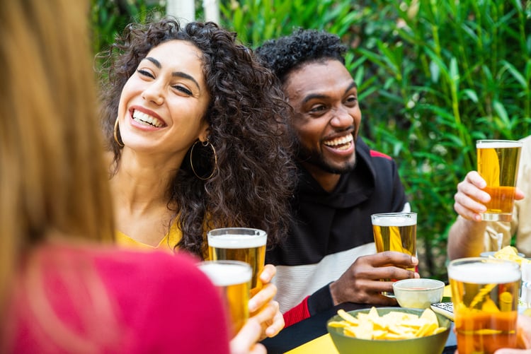 two friends smile and hold their beers while they sit outside