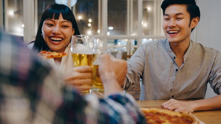 two friends smile at a table while holding pizza and beer