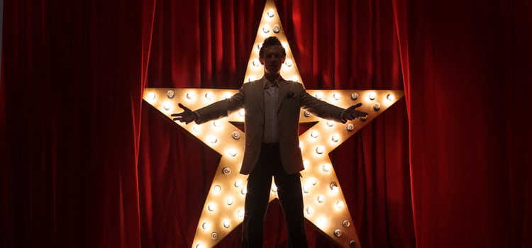 a performer stands with arms out in front of an illuminated star against a red curtain