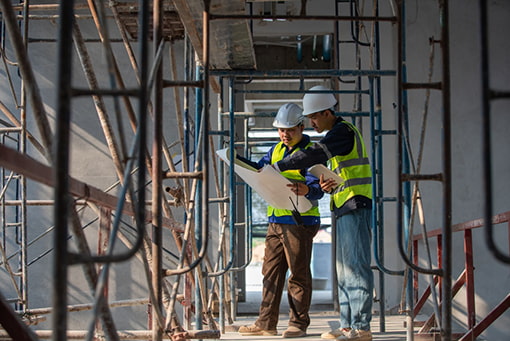 two workers in hi-vis vests look at a site plan under scaffolding